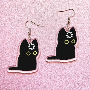 Op de afbeelding: Een paar bungelende oorbellen met een zwart kattensilhouet met gele ogen en een wit bloemmotief op het voorhoofd. De kattenbedels zijn gemaakt van doorzichtig acryl en hangen aan zilverkleurige metalen haken voor gaatjes in de oren.