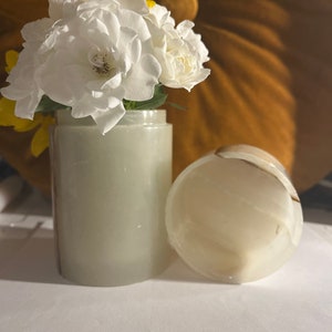 May include: A white marble vase with a white and brown marble lid. The vase is filled with white and yellow flowers.
