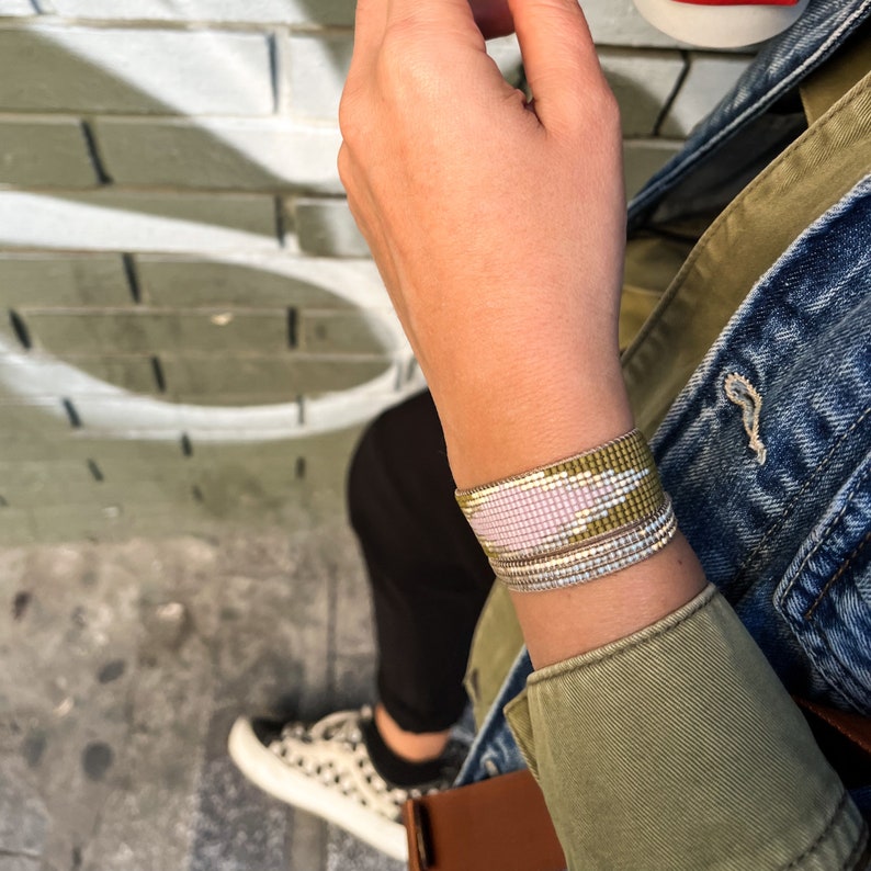 May include: A close-up of a person's wrist wearing a multi-layered beaded bracelet. The bracelet is made of small, colorful beads in shades of pink, white, and green. The bracelet is worn on a bare wrist, and the person's hand is visible in the background.
