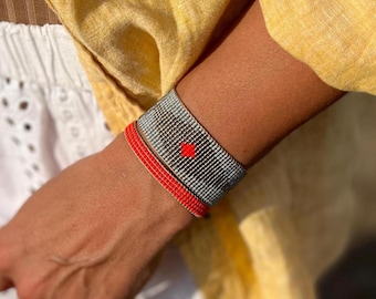 May include: Two beaded bracelets on a wrist. One bracelet is gray and white with a red square design. The other bracelet is red.