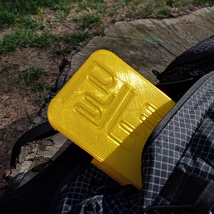 May include: A bright yellow, rectangular, 3D printed object with a textured surface. The object is partially inside a black backpack with a zipper.