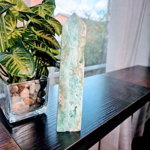 May include: A tall, rectangular crystal obelisk with a pointed top. The crystal features a blend of light blue and beige hues, with visible veining and a natural, textured surface. The obelisk is displayed on a dark wooden surface, with a potted plant in the background.