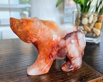 May include: A carved bear figurine made of orange and white crystal. The bear is standing on all fours, with detailed carving of its fur and features. The crystal has natural red and white veining throughout.