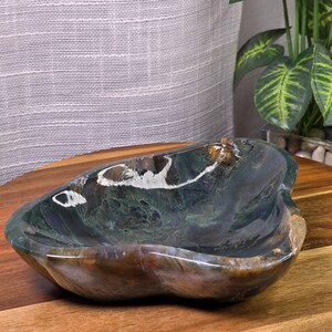 May include: A polished moss agate bowl with a smooth, organic, irregular shape. The stone features deep forest green, translucent white, and earthy brown inclusions throughout its surface, creating a natural, marbled, and multi-tonal appearance.