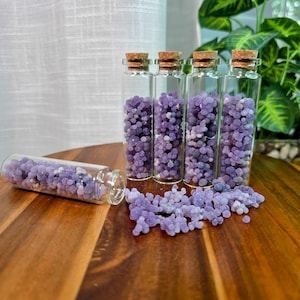 May include: Four clear glass bottles with cork stoppers, filled with small, round, purple gemstones. One bottle is lying on its side, with gemstones spilled out onto a wooden surface. The gemstones are a mix of light and dark purple.