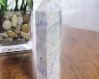 Könnte beinhalten: Eine weiße und violette Amethyst-Kristallspitze, auch bekannt als Zauberstab, wird auf einer Holzoberfläche gezeigt. Der Kristall ist facettiert und hat eine spitze Spitze.