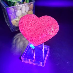 May include: A vibrant pink heart-shaped object, possibly a decorative stone or sculpture, is displayed on a clear acrylic stand. The heart has a textured surface and is illuminated by a blue light, creating a striking visual effect. The object is a unique decorative item.