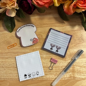 以下が含まれることがあります： ピンクのクリップ、黒いペン、小さなオレンジ色のクリップが付いた、4種類の異なる模様の付箋パッドのセット。付箋パッドには、異なるイラストと「Pure Bread Good Boy」と「The Study Goblin」のテキストが描かれています。