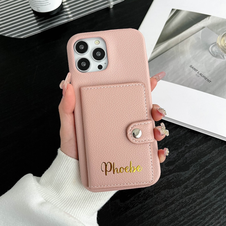 Op de afbeelding: Een roze leren telefoonhoesje met een ingebouwd portemonnee en een gouden inscriptie die "Phoebe" luidt.
