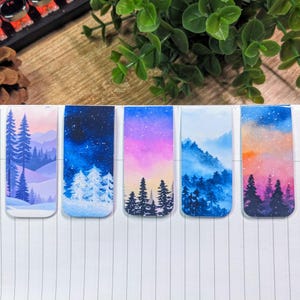 May include: Five watercolour painted magnetic bookmarks with wintery scenes. Each bookmark features a different landscape with trees, mountains, and a sky with stars, clouds, or a sunset.