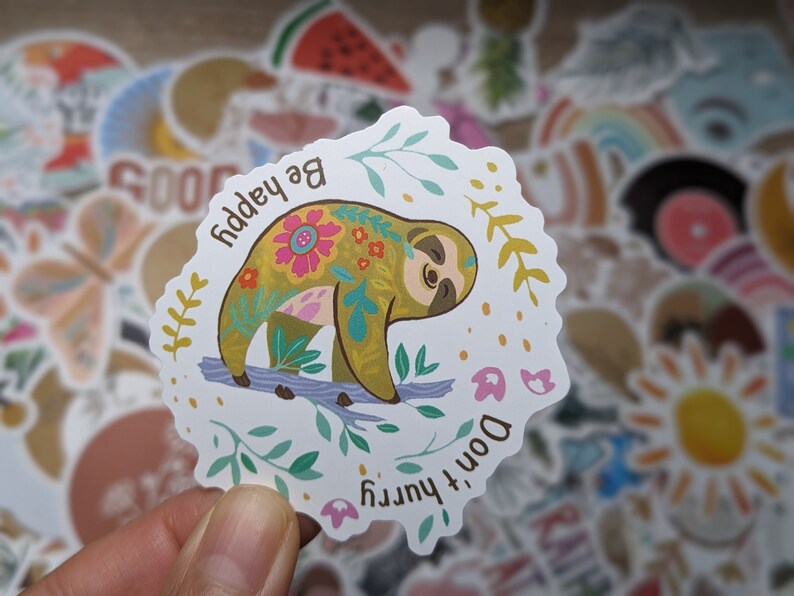 May include: A white sticker with a colorful sloth on a branch. The sloth is decorated with floral patterns and the text "Be happy, Don't hurry" is written around it.
