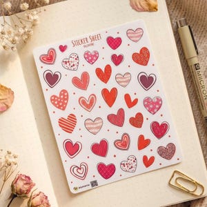 May include: A white sticker sheet featuring various heart designs in shades of red, pink, and maroon. The hearts display diverse patterns including stripes, dots, and swirls. The text 'STICKER SHEET VALENTINE' is printed at the top centre.