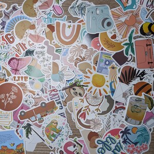 May include: A collection of colorful stickers featuring various designs, including flowers, animals, and abstract patterns. The stickers are arranged in a random pattern on a wooden surface. Some of the stickers have text, such as "VIFE", "TOMORROW", and "Be happy".