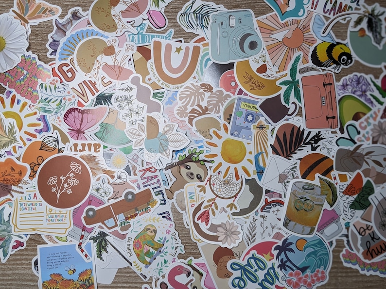 May include: A collection of colorful stickers featuring various designs, including flowers, animals, and abstract patterns. The stickers are arranged in a random pattern on a wooden surface. Some of the stickers have text, such as "VIFE", "TOMORROW", and "Be happy".