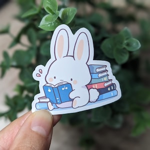 May include: A white bunny sticker reading a blue book, surrounded by stacks of colorful books. The bunny has pink inner ears and is sweating. The sticker is on a white background.