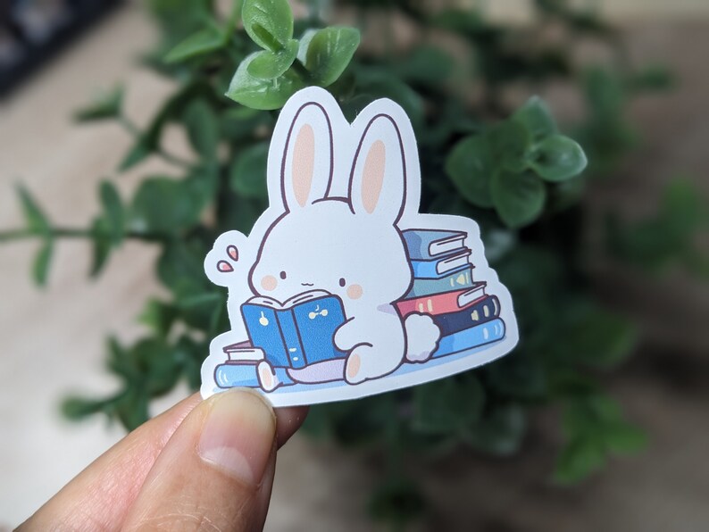 May include: A white bunny sticker reading a blue book, surrounded by stacks of colorful books. The bunny has pink inner ears and is sweating. The sticker is on a white background.