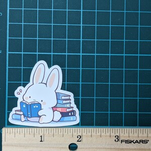 May include: A white bunny sticker reading a blue book, surrounded by a stack of books. The sticker is approximately 2 inches wide and 2 inches tall. The bunny has pink inner ears and cheeks. The books are blue and red.