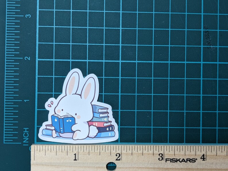May include: A white bunny sticker reading a blue book, surrounded by a stack of books. The sticker is approximately 2 inches wide and 2 inches tall. The bunny has pink inner ears and cheeks. The books are blue and red.