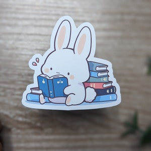 May include: A white bunny sticker reading a blue book, surrounded by a stack of colorful books. The bunny has pink cheeks and is sitting down. The sticker is on a white background.
