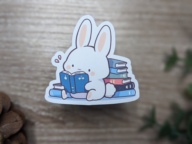May include: A white bunny sticker reading a blue book, surrounded by a stack of colorful books. The bunny has pink cheeks and is sitting down. The sticker is on a white background.