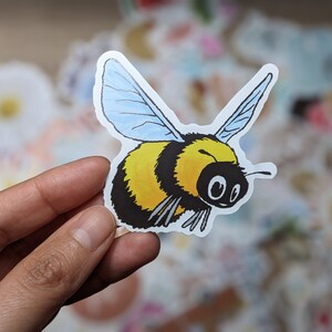 May include: A yellow and black bumblebee sticker with blue wings. The bumblebee is flying with its wings spread out. The sticker is on a white background.