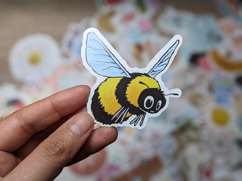 May include: A yellow and black bumblebee sticker with blue wings. The bumblebee is flying with its wings spread out. The sticker is on a white background.