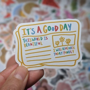 May include: A colorful sticker with a newspaper design. The headline reads "It's a Good Day". The text below the headline says "This world is beautiful" and "I will remember today fondly".