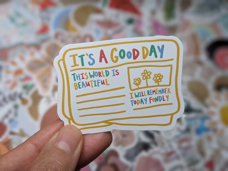 May include: A colorful sticker with a newspaper design. The headline reads "It's a Good Day". The text below the headline says "This world is beautiful" and "I will remember today fondly".