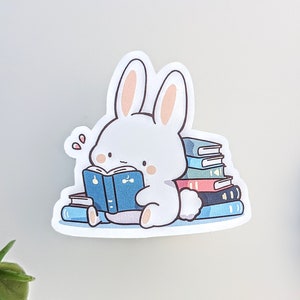 May include: A white bunny sticker reading a blue book, surrounded by a stack of colorful books. The bunny has pink inner ears and blush. The sticker has a cute, cartoonish style, perfect for book lovers and stationery enthusiasts.