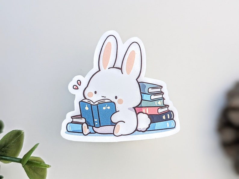 May include: A white bunny sticker reading a blue book, surrounded by a stack of colorful books. The bunny has pink inner ears and blush. The sticker has a cute, cartoonish style, perfect for book lovers and stationery enthusiasts.