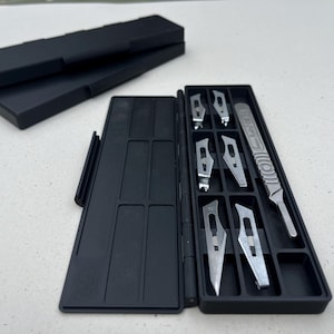 May include: A black plastic storage case containing a metal scalpel handle and six interchangeable silver scalpel blades. The case is rectangular with a hinged lid, featuring dedicated compartments for the handle and blades. The handle has text.