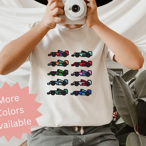 Könnte beinhalten: Weißes T-Shirt mit einer bunten Grafik von mehreren Rennwagen. Der Text "Mehr Farben verfügbar" befindet sich auf einem rosa Stern.