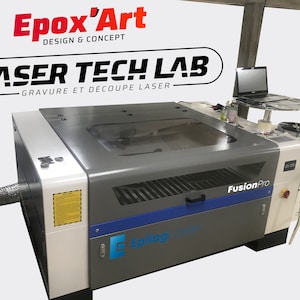 Op de afbeelding: Een witte en grijze lasercutter met een doorzichtig deksel, met de tekst "FusionPro" en "Epilog Laser". De machine staat in een werkplaats met een computerscherm en een kruk.