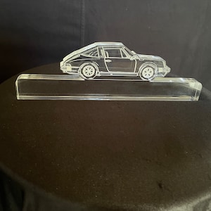 Puede incluir: Una escultura de acrílico transparente de un coche clásico, posiblemente un Porsche, sobre una base de acrílico transparente. La escultura es una representación detallada del coche, con ruedas, ventanas y líneas de carrocería visibles.