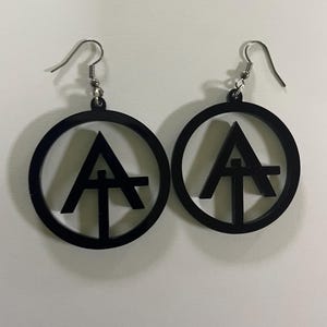 Puede incluir: Un par de pendientes negros colgantes con un diseño circular que presenta un símbolo de triángulo estilizado en su interior. Los pendientes tienen ganchos plateados.