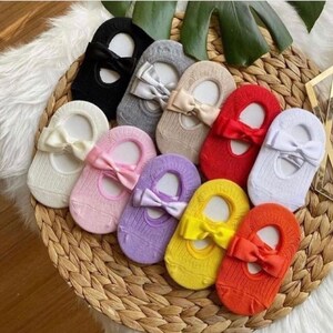 K&ouml;nnte beinhalten: Eine Sammlung bunter F&uuml;&szlig;linge mit Schleifenakzenten. Die Socken sind in verschiedenen Farben erh&auml;ltlich, darunter Schwarz, Grau, Beige, Rot, Wei&szlig;, Rosa, Lila, Gelb und Orange. Sie sind auf einem geflochtenen Weidenkorb angeordnet.