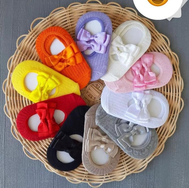 K&ouml;nnte beinhalten: Eine Sammlung bunter F&uuml;&szlig;linge mit dekorativen Schleifen. Die Socken sind in verschiedenen Farben erh&auml;ltlich, darunter Orange, Gelb, Rot, Schwarz, Grau, Beige, Wei&szlig;, Rosa und Lila. Sie sind in einem Weidenkorb angeordnet.