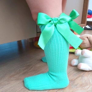 May include: A pair of light teal knee-high socks with a large, bright green bow at the top. The socks are ribbed and appear to be made of a soft material. The bow adds a decorative touch to the socks.