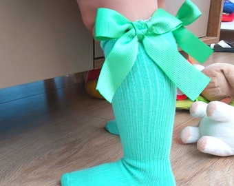 May include: Light teal knee-high socks with a large, bright green bow. The socks have a ribbed texture and are made of a soft material. The bow is a decorative feature. The socks are suitable for children.