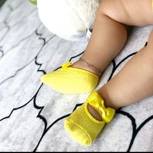 K&ouml;nnte beinhalten: Ein Paar hellgelbe Babysocken mit Schleifendetail. Die Socken sind gerippt und haben einen Mary-Jane-Stil. Die Beine des Babys sind sichtbar, mit einem Fu&szlig;kettchen. Die Socken liegen auf einer gemusterten Decke.