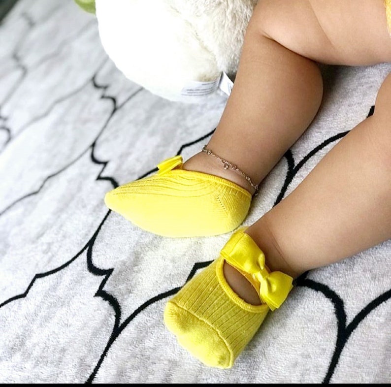 K&ouml;nnte beinhalten: Ein Paar hellgelbe Babysocken mit Schleifendetail. Die Socken sind gerippt und haben einen Mary-Jane-Stil. Die Beine des Babys sind sichtbar, mit einem Fu&szlig;kettchen. Die Socken liegen auf einer gemusterten Decke.
