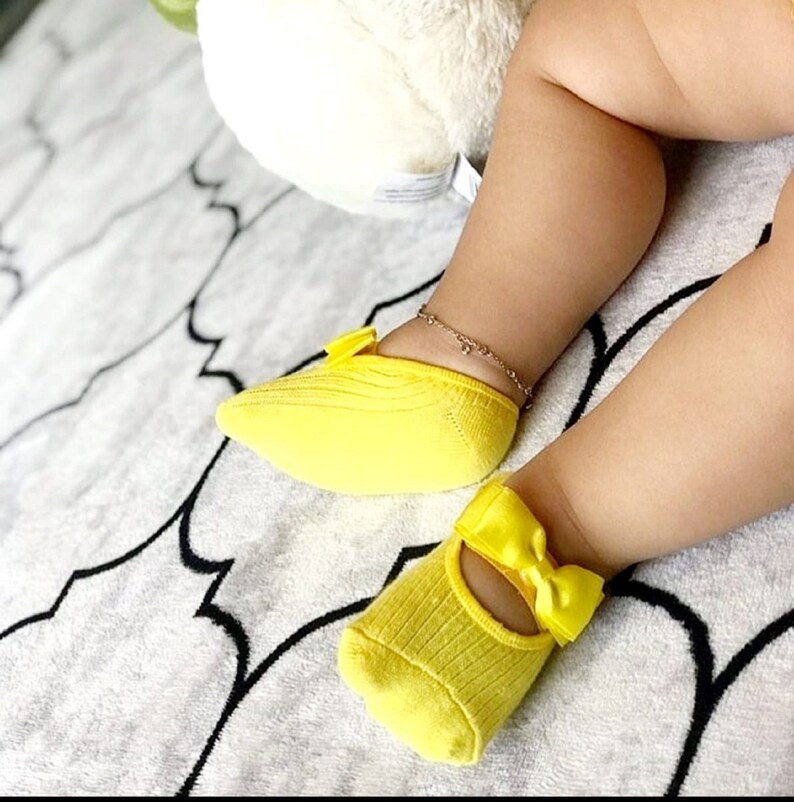 K&ouml;nnte beinhalten: Ein Paar hellgelbe Babysocken mit Schleifenakzenten. Die Socken sind gerippt und haben einen Mary-Jane-Stil mit einer Schleife oben. Am Kn&ouml;chel des Babys befindet sich eine zarte goldene Fu&szlig;kette. Die Socken liegen auf einer wei&szlig;-schwarz gemusterten Decke.