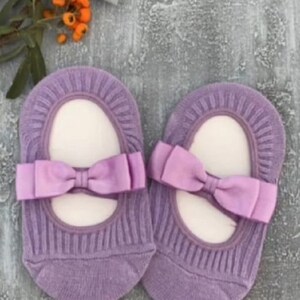 K&ouml;nnte beinhalten: Ein Paar lavendelfarbene Babysocken mit Schleifendetail. Die Socken haben eine gerippte Textur und eine Satinschleife in passendem Lavendelton. Die Socken sind so gestaltet, dass sie Ballerinas &auml;hneln.