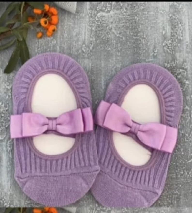 K&ouml;nnte beinhalten: Ein Paar lavendelfarbene Babysocken mit Schleifendetail. Die Socken haben eine gerippte Textur und eine Satinschleife in passendem Lavendelton. Die Socken sind so gestaltet, dass sie Ballerinas &auml;hneln.