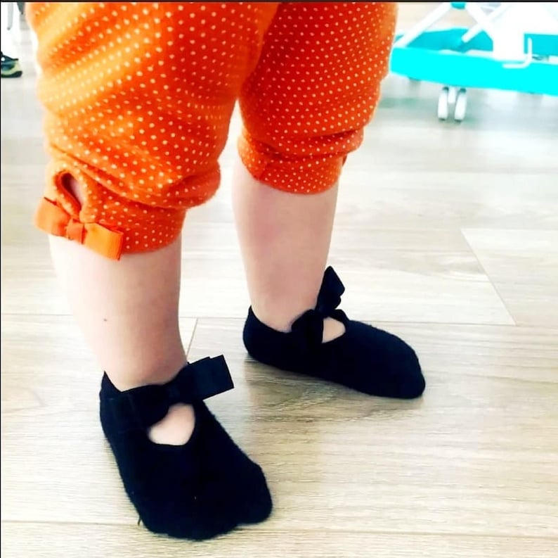 May include: A pair of black baby shoes with a bow on the front. The shoes are made of a soft, stretchy material and have a comfortable fit.
