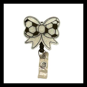 May include: A badge reel with a soccer ball bow design. The bow is white with silver glitter and black soccer ball details. The reel has a black bead, a silver clip, and a clear plastic strap.