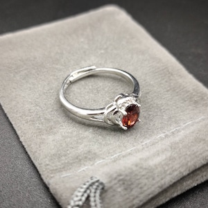 May include: A silver ring featuring a red gemstone set in an ornate design. The ring is presented on a grey velvet pouch.