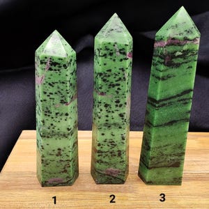 May include: Three green and pink zoisite crystal towers with pointed tops. The crystals have a speckled appearance with dark green and black spots. The crystals are displayed on a wooden surface against a black background.
