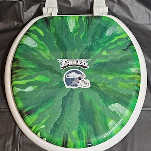 May include: Green and black toilet seat cover with a white rim. The cover has a green and black swirl pattern with a white logo that says "EAGLES" and a football helmet.