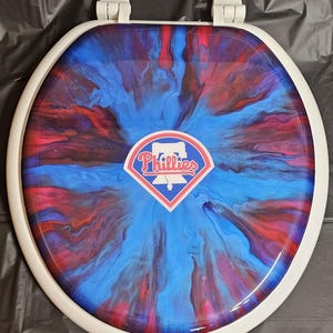 May include: White toilet seat with a blue and red abstract design. The design features a white baseball diamond with the word "Phillies" in red letters.